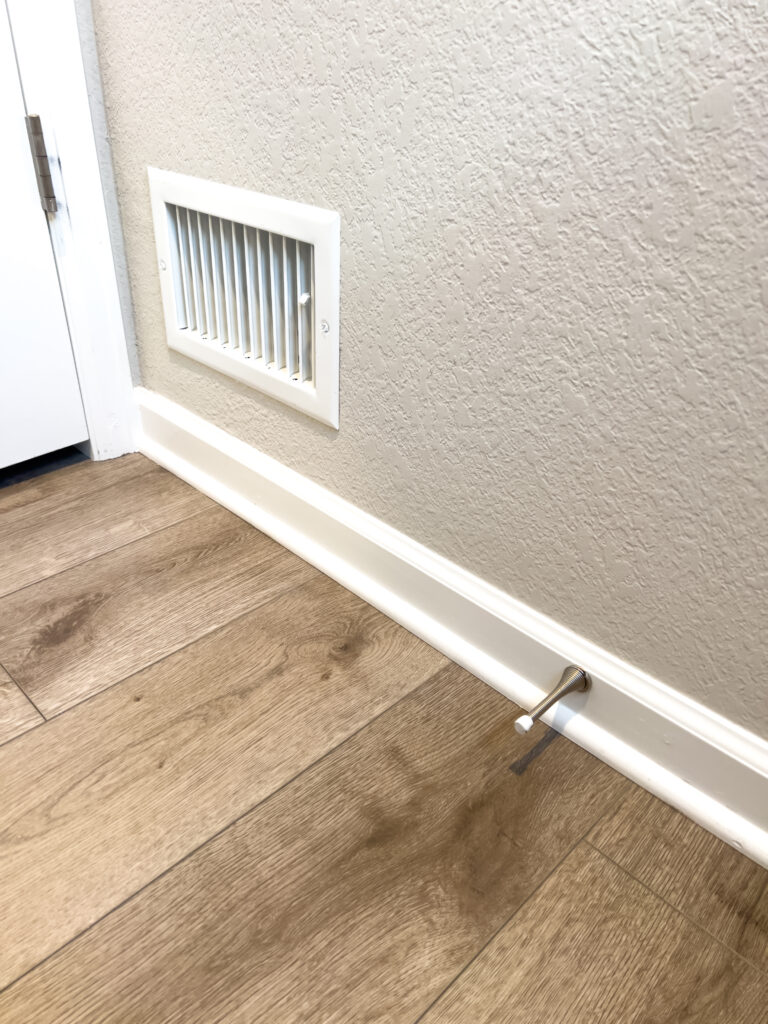 This image shows a section of a room with new wood flooring. The focus is on a white floor vent and a door stop installed on the baseboard. The vent has horizontal slats and is positioned close to the floor. The door stop is metallic and slightly protrudes from the baseboard, ready to prevent the door from hitting the wall. The new wood flooring has a light, natural finish, complementing the clean, modern look of the room.